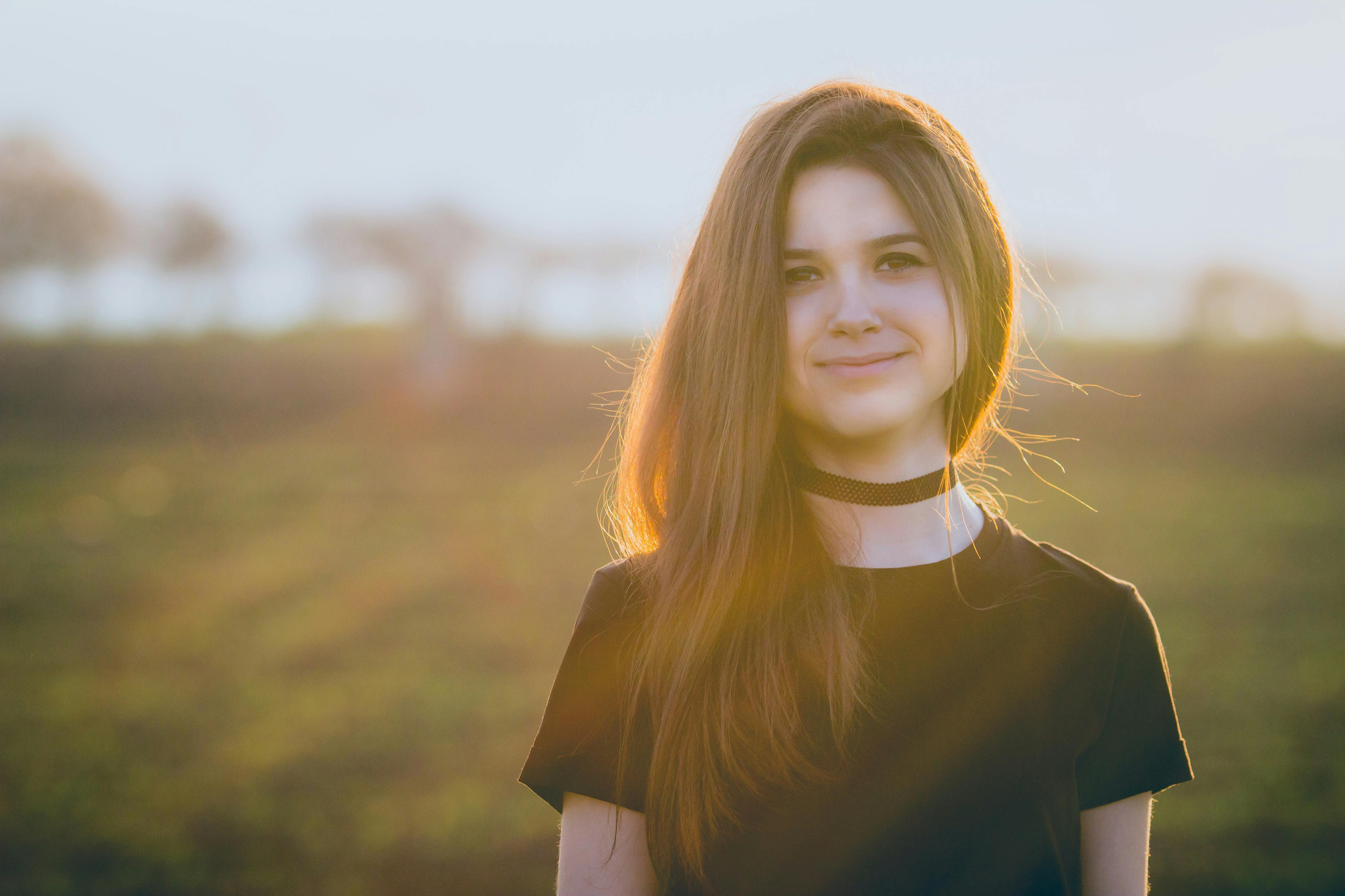A young woman stands in a field, smiling with her back to the sun, which creates a halo effect on her long brown hair.