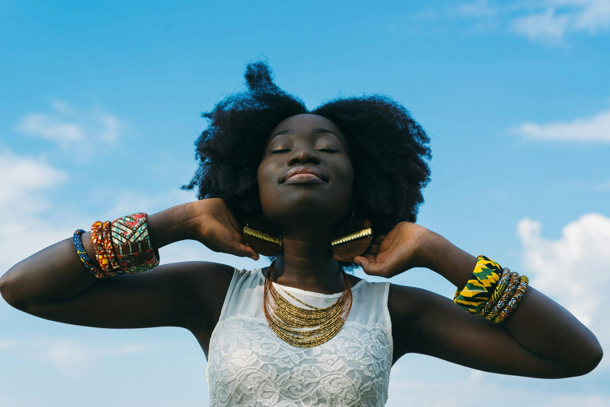 A beautiful Black woman poses with her eyes closed and chin lifted to the blue sky surrounding her, a look of peace and contentment on her face.  She wears ornately beaded tribal bracelets.