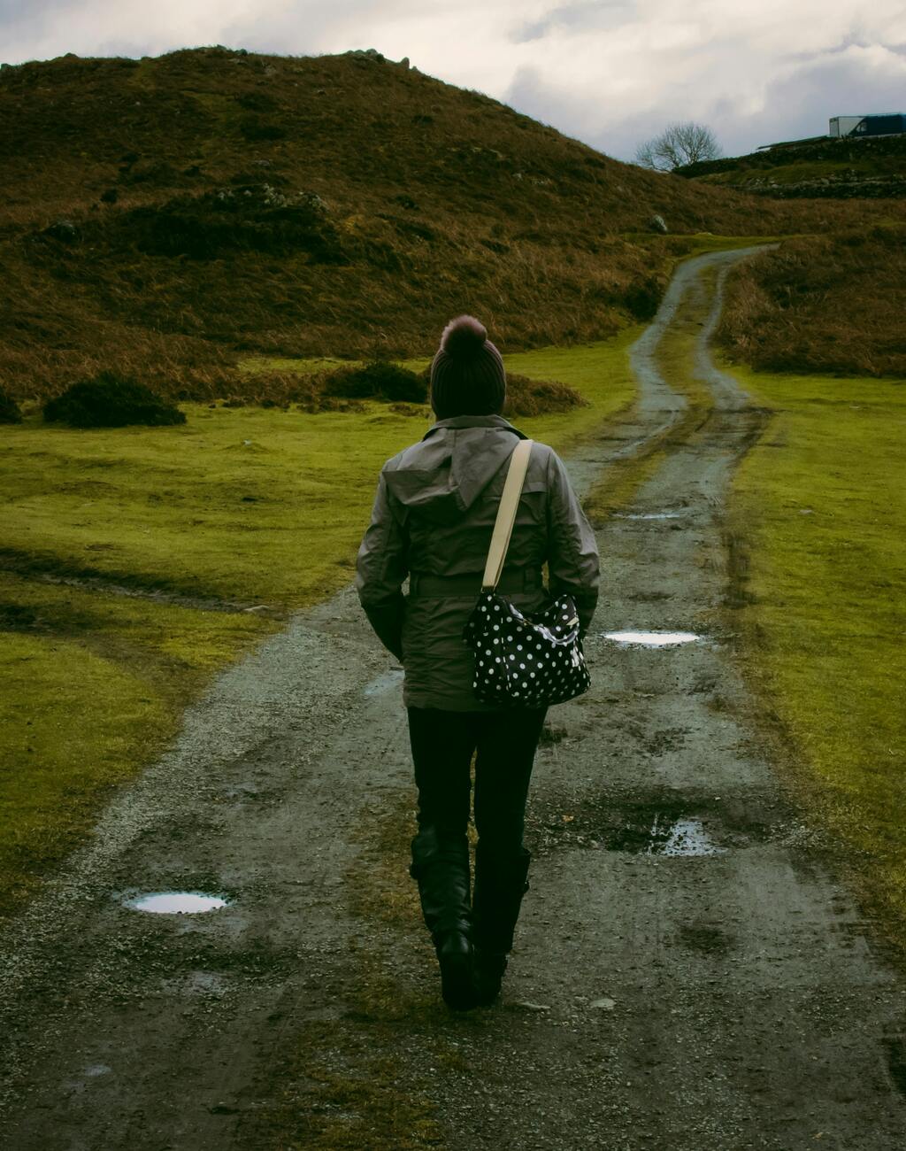 A person clad in cool weather gear and a bag across their back stands at a  fork in the road.  The landscape is hilly, green, and puddles have formed in the potholes of the dirt road.