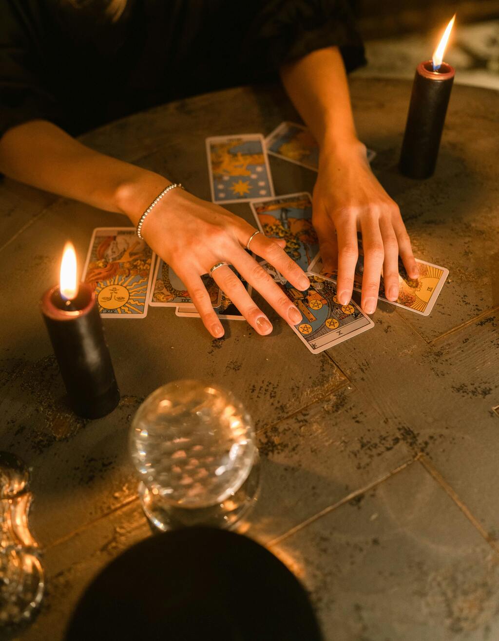 In a candle-lit room, a woman's hands can be seen spreading tarot cards out on a tiled table.  A crystal ball sits to the side.