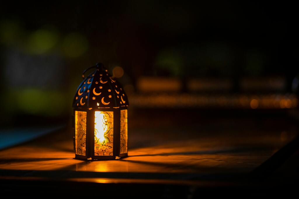 A lit metal lantern sits on top of a wooden surface, sending out rays a light into the darkness.  The lantern features cut outs of stars and crescent moons on the cap.
